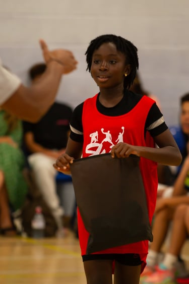 Young girl in a red basketball jersey holding a black bag at a sports camp.