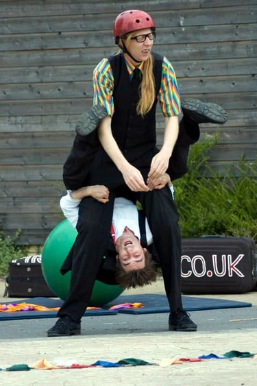 Two street performers in colorful costumes demonstrate a vertical acrobatic juggling balancing act. 