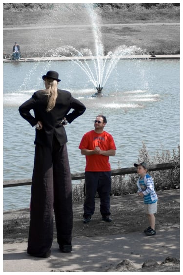 Street performer on tall stilts entertains a man and child by a park fountain.