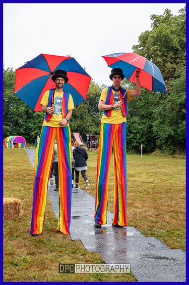 Two circus performers on stilts wearing rainbow striped trousers and top hats holding umbrellas.
