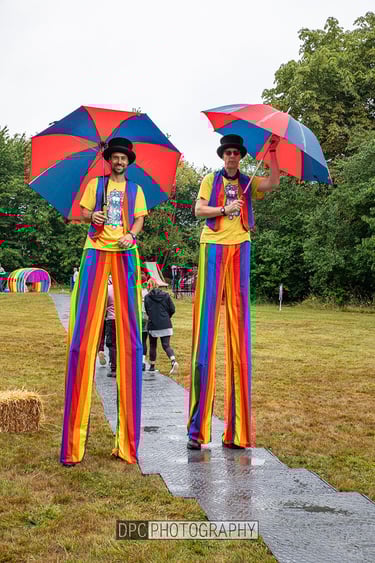 Two stilt walkers in rainbow striped trousers and top hats hold colorful umbrellas at an outdoor festival.