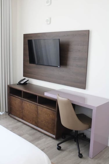 Modern hotel room with wall-mounted flat screen TV, wooden media console, and built-in work desk with ergonomic chair.