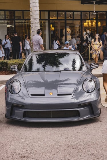 Front view of a grey Porsche 911 GT3 parked outside a high-end shopping center with palm tree reflections.