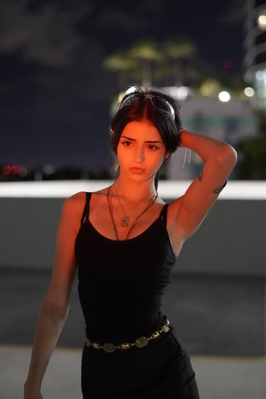 A young woman in a black tank top and gold jewelry posing at night with orange city light highlights.