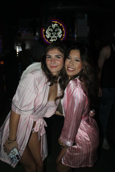 Two smiling women wearing pink striped satin robes and angel wings at a nightclub party.
