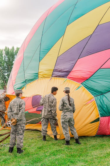 Military cadets in camouflage uniforms preparing a colorful hot air balloon for flight on a grassy field.