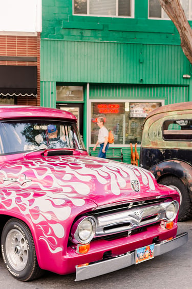 Vintage pink Ford F100 truck with white flame decals parked on a city street near storefronts.