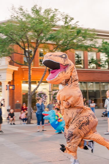 A person wearing a funny inflatable T-Rex dinosaur costume running down a city street during a parade.