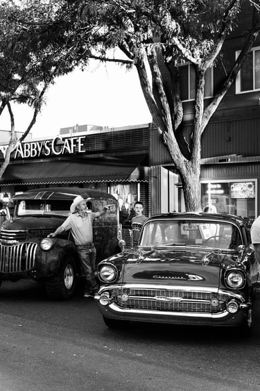 Black and white photo of a cowboy with a vintage Chevrolet car and truck outside Abby's Cafe.