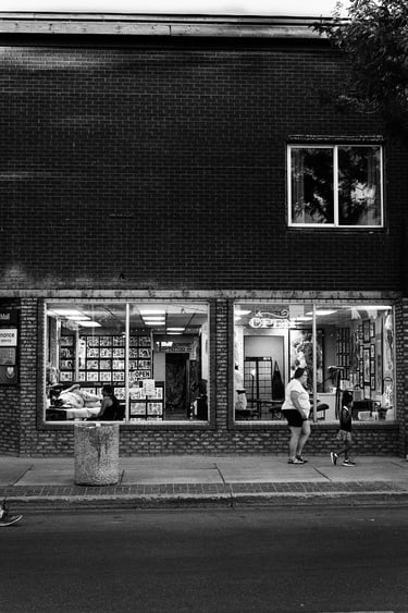 Black and white street photography of people walking past a brick tattoo parlor storefront window.
