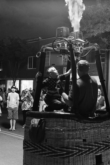 A young boy sits in a wicker hot air balloon basket at night as a pilot helps him ignite the gas burner.