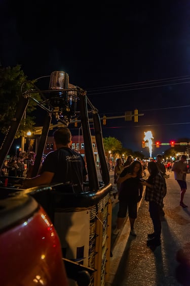 Hot air balloon burner glowing at a night festival with crowds on a city street.