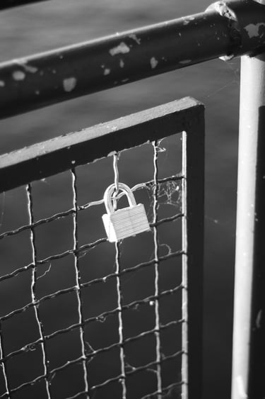 Black and white photo of a metal love lock attached to a wire mesh bridge railing.