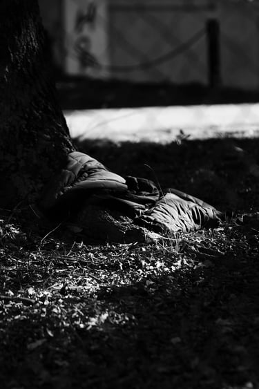 Black and white photo of a discarded puffer jacket lying on the ground at the base of a tree trunk.