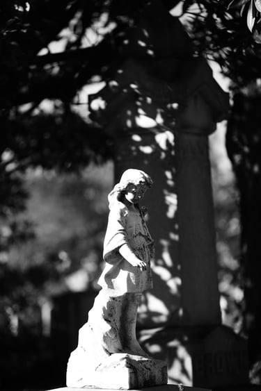 Black and white vertical photo of a stone child statue at a historic cemetery in sunlight.