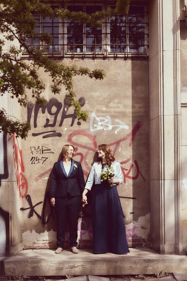Zwei Bräute halten sich an den Händen und stehen neben einer Wand mit Graffiti.