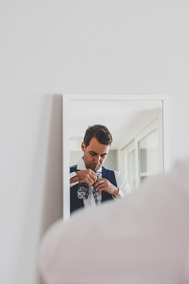 Groom getting ready, tying his tie in front of a mirror.