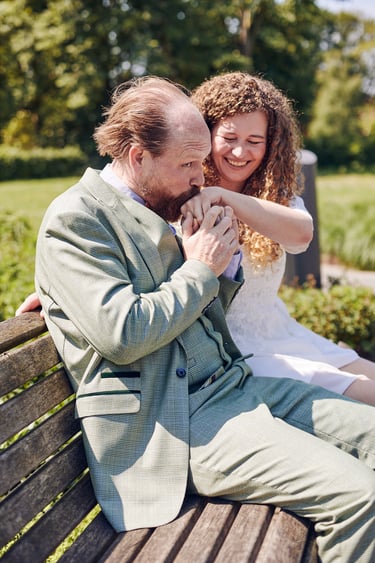 Groom kissing brides hand while sitting on a park bench.