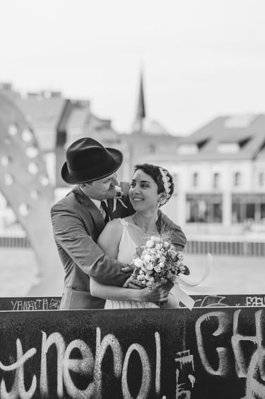 Groom with a hat hugging bride from behind and smiling at each other.