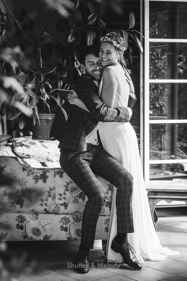 Stehende Braut umarmt sitzenden Bräutigam in einem Wintergarten.