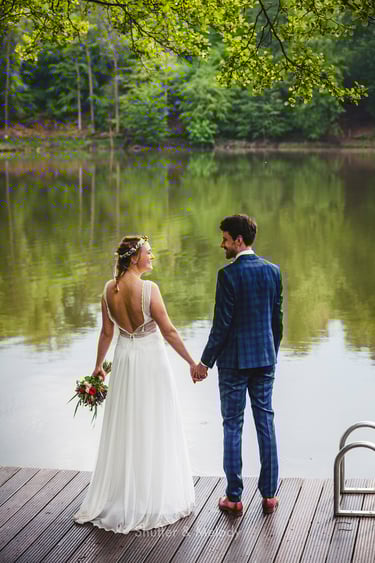 Braut und Bräutigam stehen Hand in Hand auf einem Steg neben einem See.