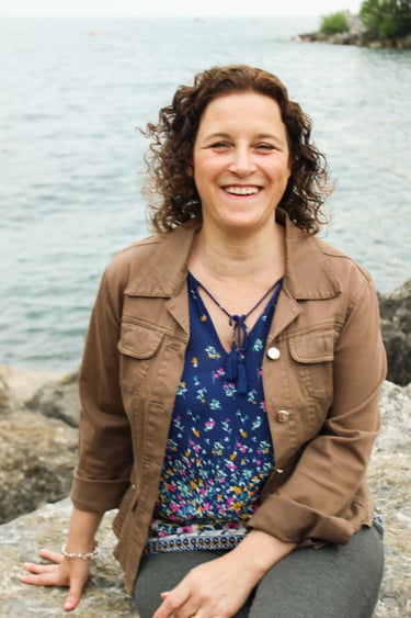 Smiling woman with curly hair sitting on rocks by a lake wearing a brown jacket and floral top.