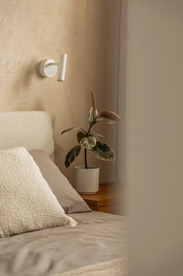 Biały kinkiet do czytania nad łóżkiem i roślina na szafce nocnej w sypialni.