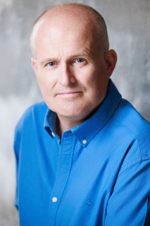 Professional headshot of a middle-aged bald man wearing a blue button-down shirt against a grey background.