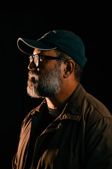Side profile of a middle-aged man with a gray beard wearing a baseball cap and glasses against a dark background.