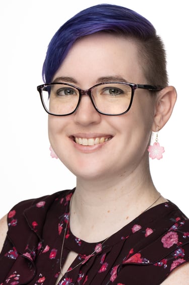 Smiling woman with short purple hair and glasses wearing floral pink earrings and a dark blouse.