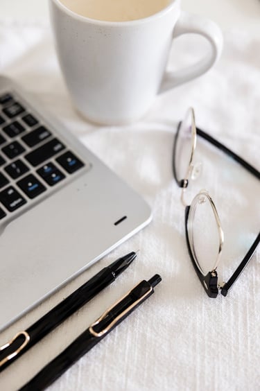A laptop, pens, reading glasses, and coffee cup on a white table