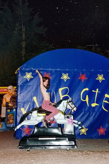 A woman in striped pants riding a vintage coin-operated horse ride at a carnival at night.
