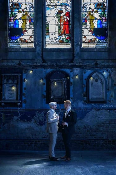 Groom and guest talk inside a rustic wedding venue with stained glass windows and stone walls.