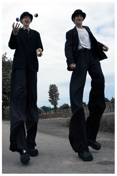 Two street performers on high stilts wearing black suits while juggling balls outdoors.
