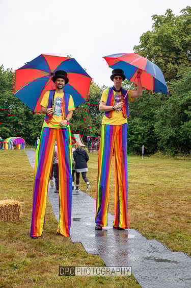 Two circus entertainers on stilts wearing rainbow trousers and top hats with umbrellas at an outdoor festival.