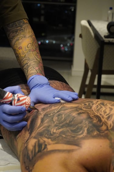 Professional tattoo artist in blue gloves using a machine to ink a detailed full back tattoo.