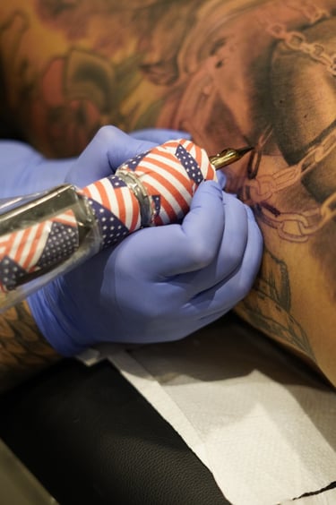 Professional tattoo artist in blue gloves using an American flag themed machine to ink a detailed design.