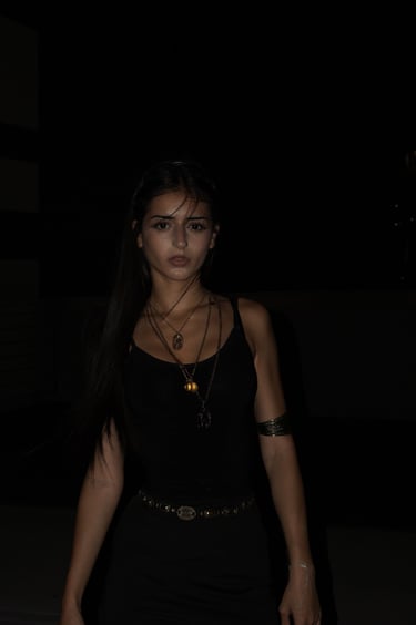 Young woman in a black tank top wearing layered gold necklaces and a bohemian belt at night.