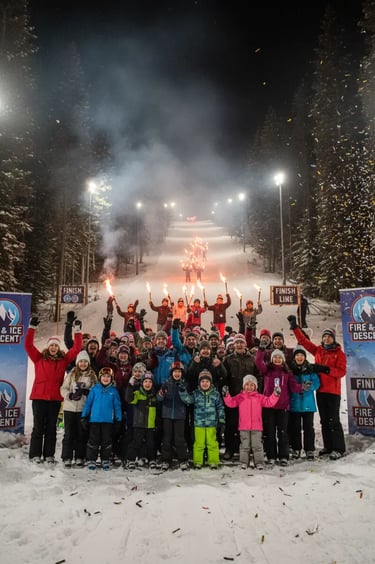 A large group of people posing for a photo on a snowy ski slope at night, holding torches with a lit
