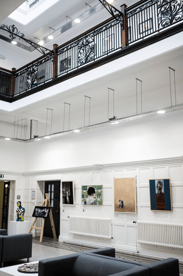 Paintings in a gallery with sofas on ground level underneath a balcony.  Partial glass ceiling. 