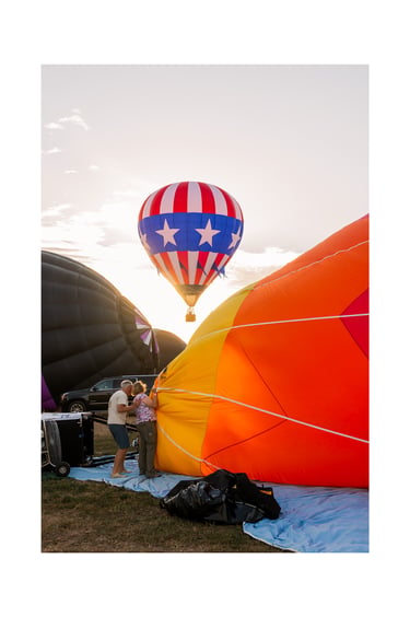 A patriotic star-patterned hot air balloon flying over a colorful festival launch field at sunrise.