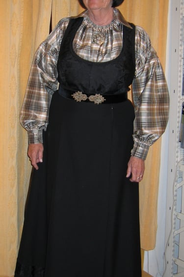 A woman with black hair and glasses stands in a black bunad with a plaid shirt