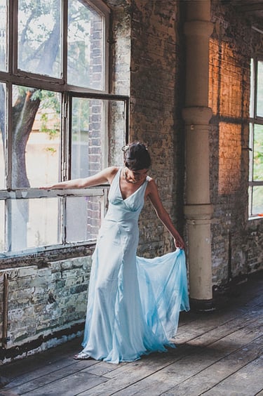 Eine Braut in einem hellblauen Kleid steht neben einem Fenster und richtet ihr Kleid.
