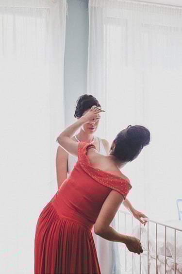 A bridesmaid in a red dress fixing bride's hair in a well lit bedroom.
