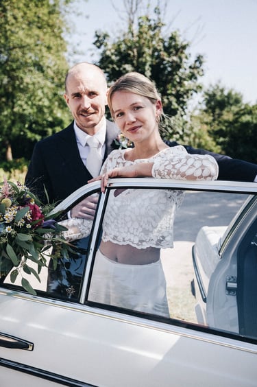 Bride and groom standing next to a car with an open door.