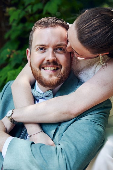 Groom looking directly into the camera as his wife hugs him from behind.