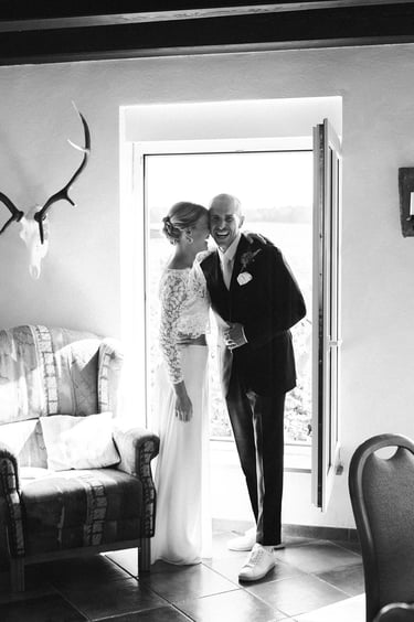 Braut und Bräutigam stehen lachend vor einem offenen Fenster.