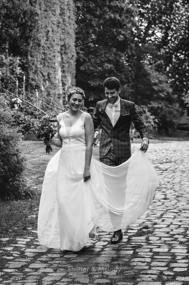 Der Bräutigam hält das Brautkleid, während sie über einen gepflasterten Weg gehen.