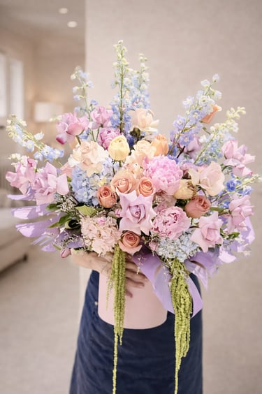 A large pastel flower arrangement featuring pink peonies, peach roses, and blue delphinium in a pink box.