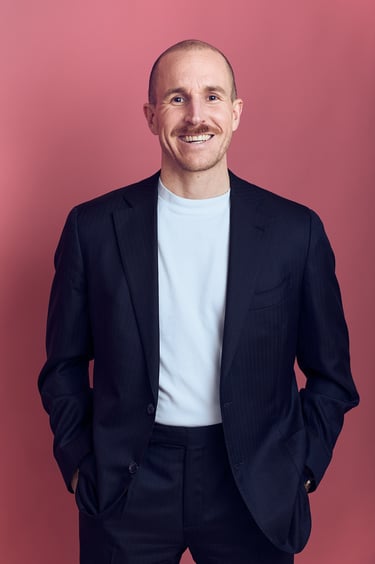 A man in a suit and a t-shirt photographed in front of a pink background.